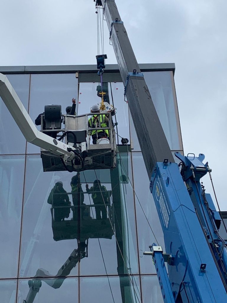 Close-up: Workers in a boom lift guide a large glass panel being lowered by a crane onto a glass building.