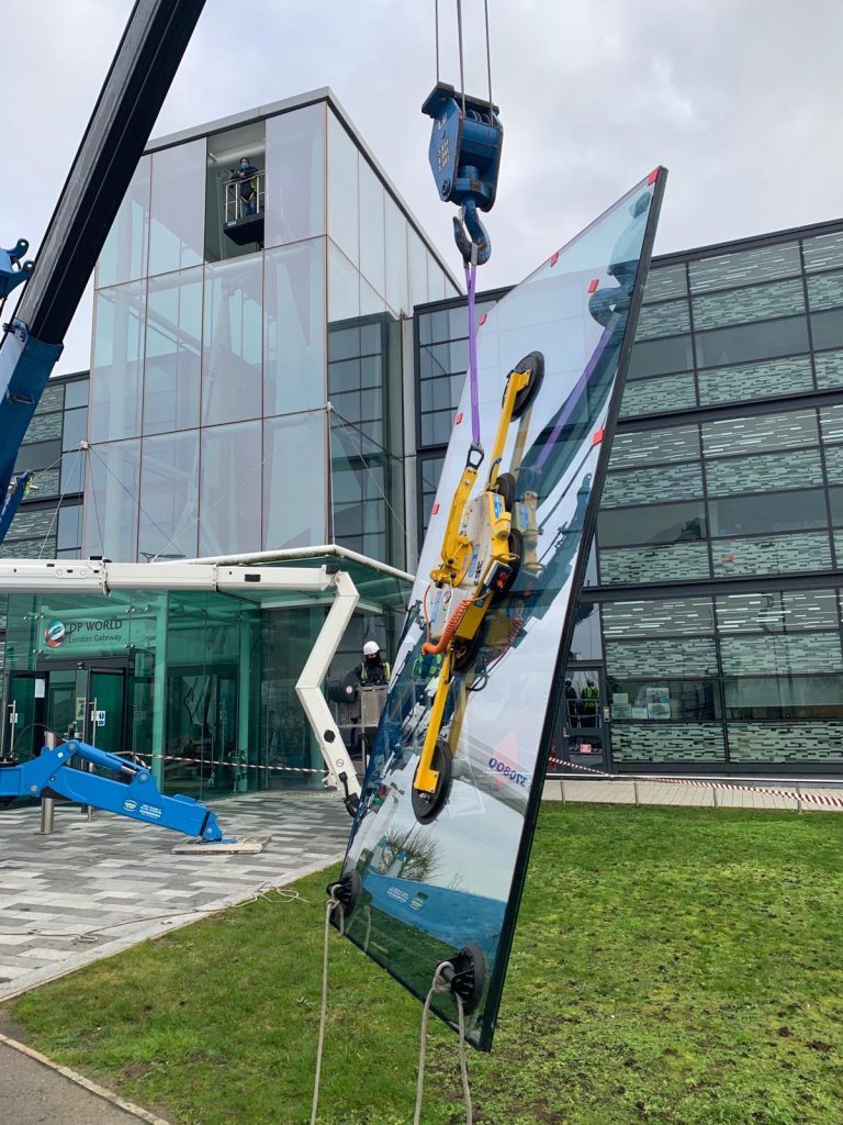 Crane lowering a large glass panel with suction cups towards a modern glass-fronted building.