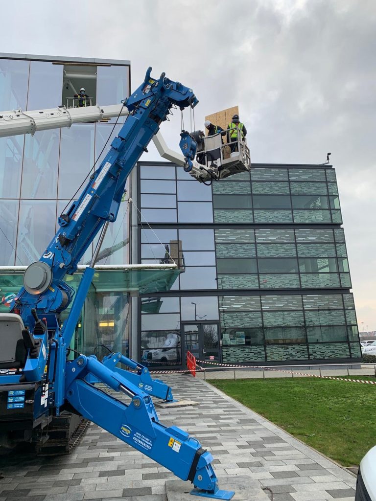 Angle shot: A blue crane and a white boom lift assist workers installing glass on a multi-story building.