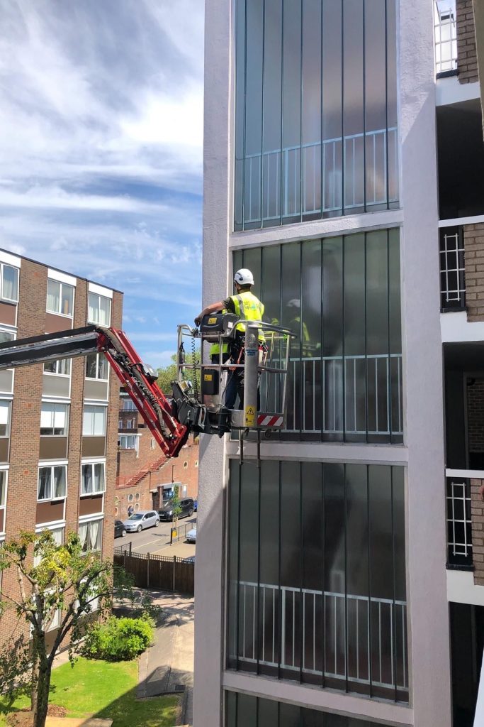 Worker on a lift installing tall aluminium framed windows on a multi-storey residential building.